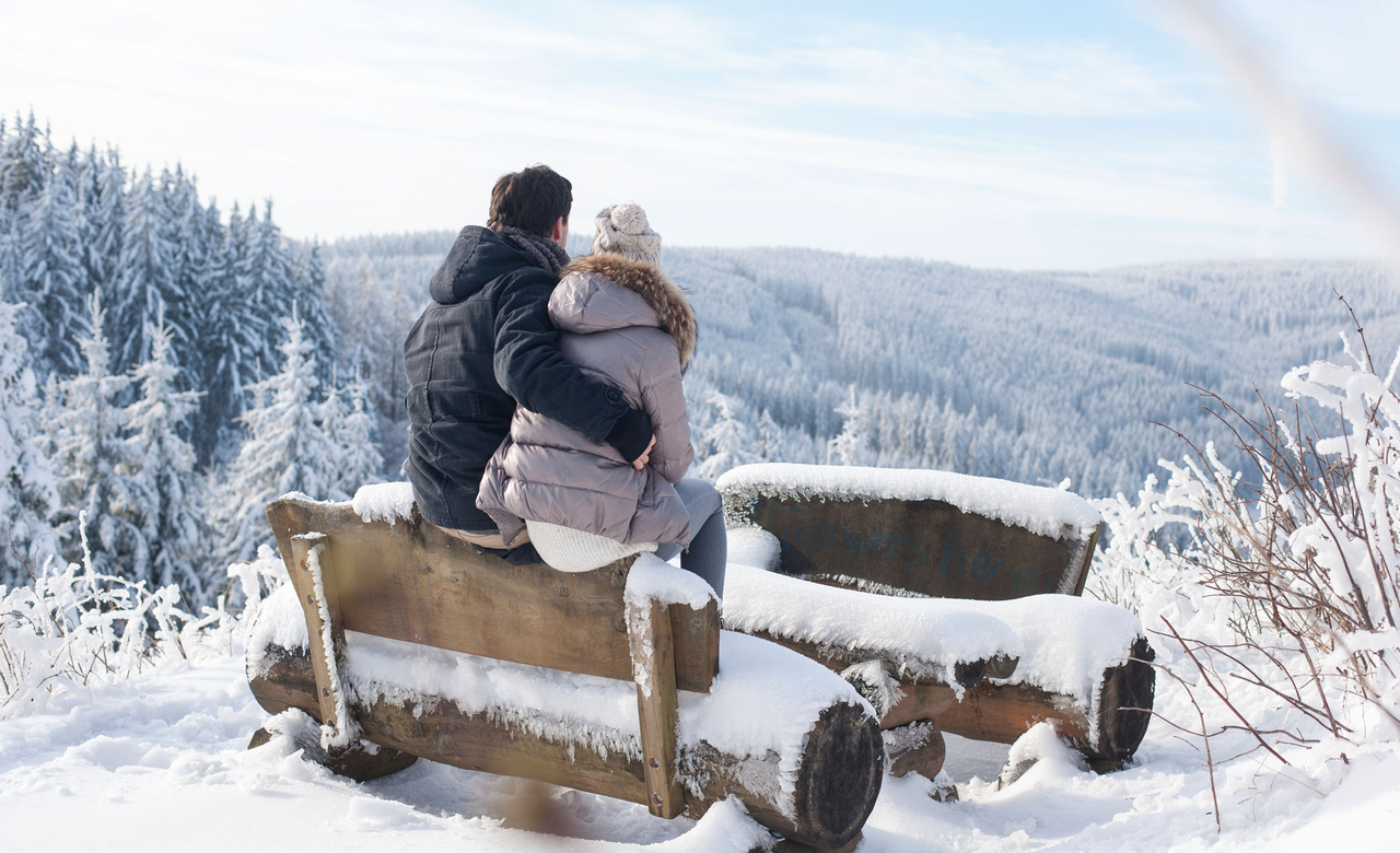 junges Paar sitzt an einer Wanderraststätte auf einer Bank und blickt auf die verschneite Winterlandschaft des Thüringer Waldes