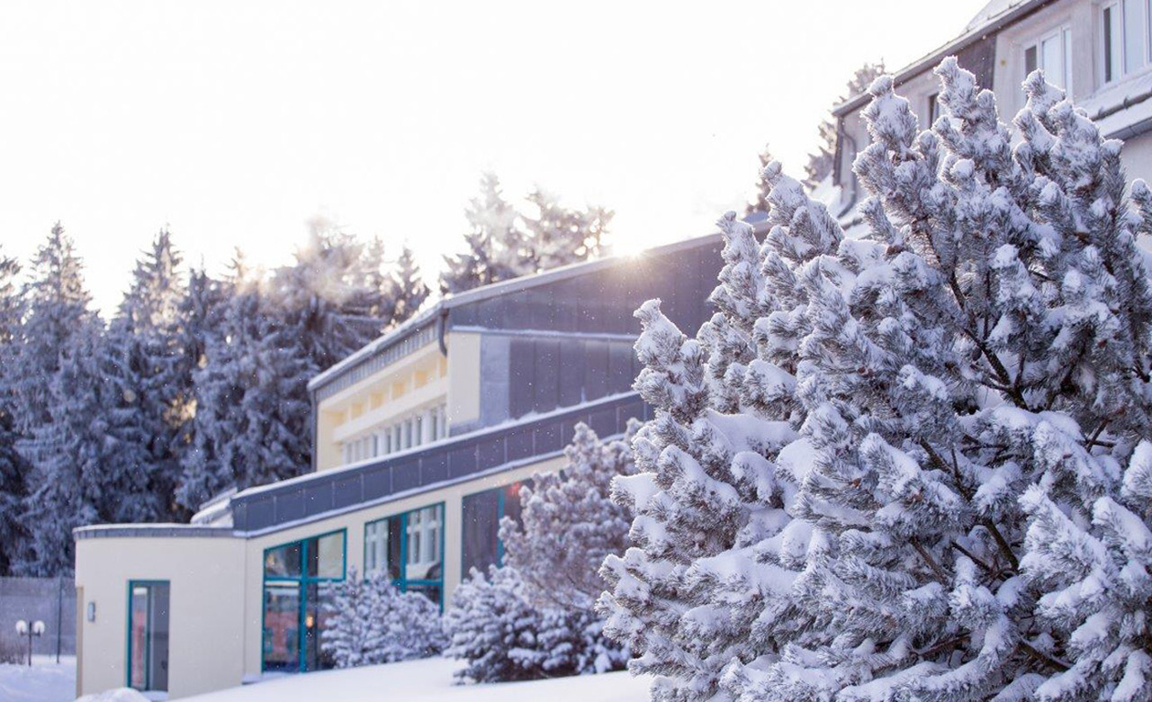 Außenansicht vom Sporthotel Oberhof mit Blick auf den Eingangsbereich eingebettet in der verschneiten Winterlandschaft des Thüringer Waldes